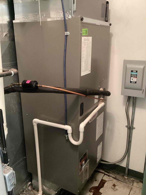 Responded to emergency service call for water leak from upstairs AC unit in commercial medical office causing damage to floor and water intrusion into mop bucket room and reception area bathroom below. Upon arrival, inspected the affected air handler unit and surrounding areas. Documented existing HVAC installation including gray metal air handler cabinet, refrigerant line connections, condensate drainage system with PVC trap configuration, and associated electrical components. Photographed the complete system setup showing copper refrigerant lines with white foam insulation, condensate drain piping, electrical disconnect box, and all mechanical connections. Identified water accumulation issues affecting the flooring beneath the unit with visible staining on concrete surface. Assessed the condensate drainage system including the U-trap configuration and drain line routing to determine source of water leak causing damage to lower level spaces. Evaluated all plumbing connections, drain p
