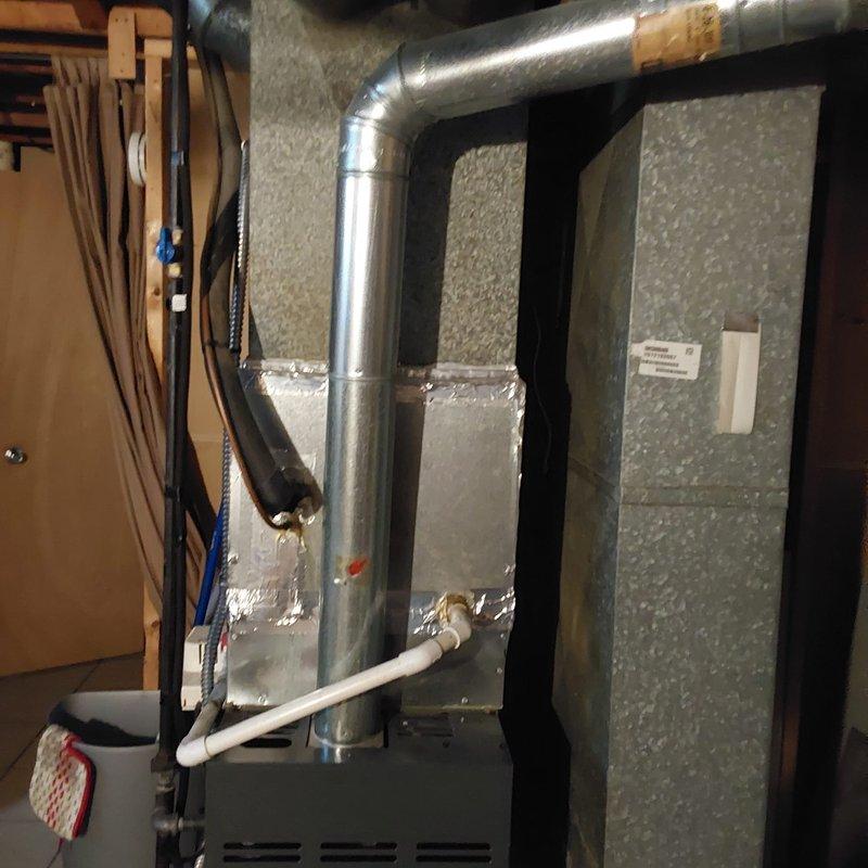 Completed full AC system retrofit including replacement of outdoor condensing unit and indoor air handler with associated ductwork modifications. Installed new gray air handler unit in basement utility area with insulated supply and return ducts, secured with foil-backed insulation and metallic tape at all seams. Connected metallic ductwork transitions from air handler to existing distribution system. Installed new outdoor condensing unit with black mesh grille and white fan cover on existing concrete pad. Unit positioned against exterior wall with proper clearance maintained. Connected electrical service through existing disconnect box with appropriate conduit routing. Installed PVC condensate drain lines with brass fittings and shutoff valves for proper drainage. All refrigerant lines properly insulated and secured between indoor and outdoor equipment.
