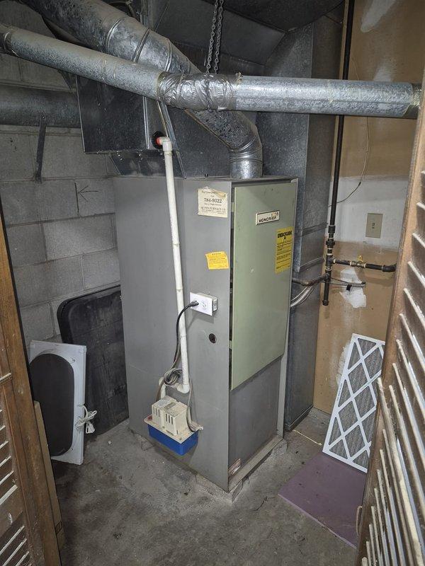 Responded to service call for air conditioning system with unknown unit age. Upon arrival, located outdoor condenser unit in covered exterior utility area showing signs of weathering and debris accumulation around base. Inspected indoor furnace and air handler system in basement utility room, noting standard equipment configuration with ductwork, refrigerant lines, and condensate management components in place. Performed diagnostic evaluation of AC system operation and documented equipment condition through photography for service records.