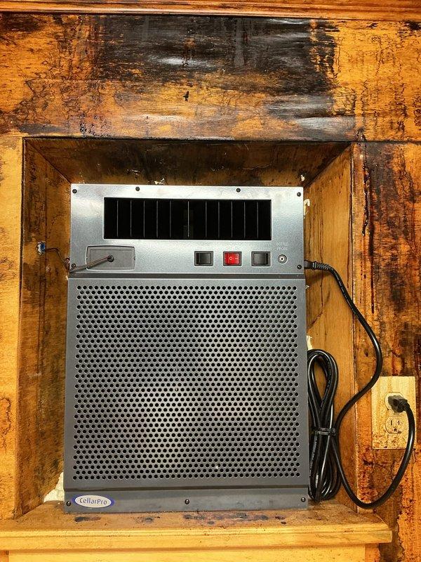 Completed installation of CellarPro through-wall wine cooling unit in wooden alcove with integrated digital thermostat control. Unit was properly mounted in recessed wooden cabinet space, electrical connection established via standard wall outlet, and Copeland digital thermostat installed on adjacent wall surface displaying operational temperature reading of 63°F with system status indicators functioning correctly.