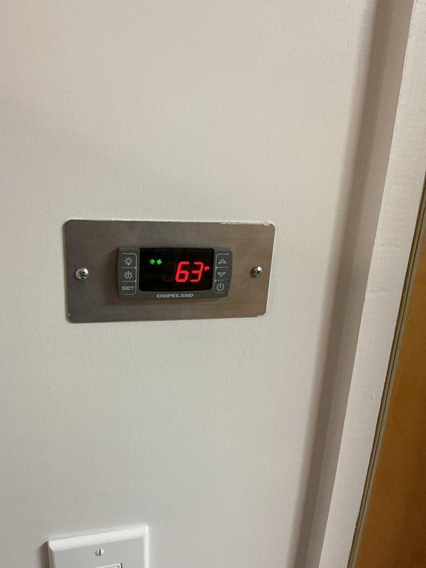 Completed installation of CellarPro through-wall wine cooling unit in wooden alcove with integrated digital thermostat control. Unit was properly mounted in recessed wooden cabinet space, electrical connection established via standard wall outlet, and Copeland digital thermostat installed on adjacent wall surface displaying operational temperature reading of 63°F with system status indicators functioning correctly.