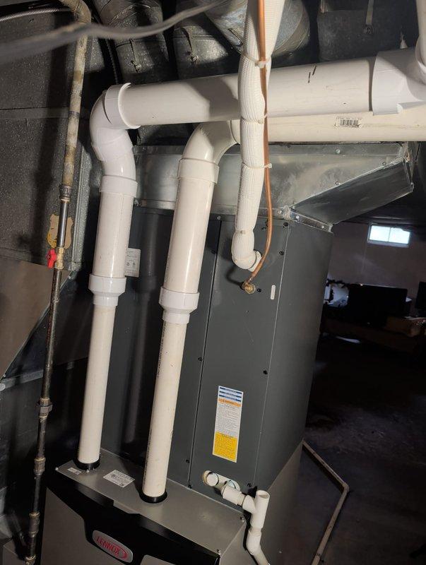 Completed furnace retrofit installation in crawl space/basement area. Installed new gray metal air handler unit with proper venting system utilizing white PVC piping secured to ceiling joists with appropriate strapping. Configured condensate drainage system with multiple PVC drain lines running from unit to designated discharge point. Connected black flexible ductwork to air handler for proper air distribution throughout structure. Verified all manufacturer specifications and safety requirements met, with proper labeling visible on equipment. Installation includes condensate pump mounted at base of unit for efficient moisture removal. All piping connections sealed and tested for leaks, with copper and gas lines properly routed along ceiling joists per code requirements.