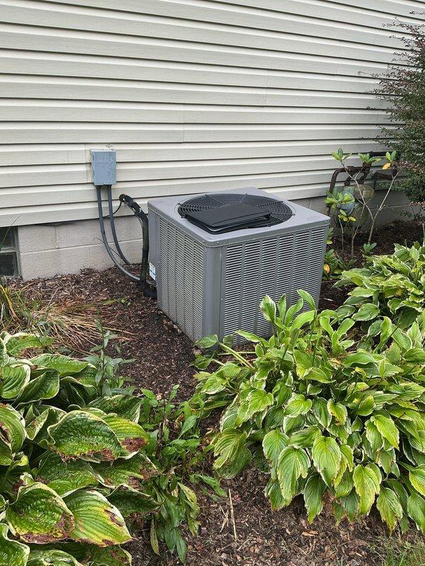 Completed full air conditioning system replacement at residential property. Installed new outdoor condenser unit on existing concrete pad with proper clearances maintained from surrounding landscaping and hostas. Mounted new electrical disconnect box on exterior wall with code-compliant conduit routing from house to unit. Interior installation included new air handler positioned in basement mechanical room alongside existing furnace and water heater in confined utility space. Routed new white PVC condensate drain lines vertically with proper pitch and termination. All refrigerant lines connected and pressure tested, system charged to manufacturer specifications. Verified electrical connections at both indoor and outdoor units, tested all safety controls and disconnect functionality. System startup completed with airflow measurements confirming proper CFM delivery. Verified cooling operation through complete cycle, checked temperature split across coil within acceptable range. All equip