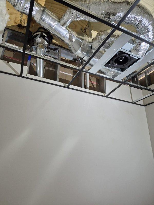 Completed installation and finishing of exhaust fan system with flexible metallic ductwork routing through exposed ceiling cavity. Installed ceiling-mounted exhaust grille with integrated fan component and wall-mounted return air vent, ensuring proper ventilation for the new construction space.