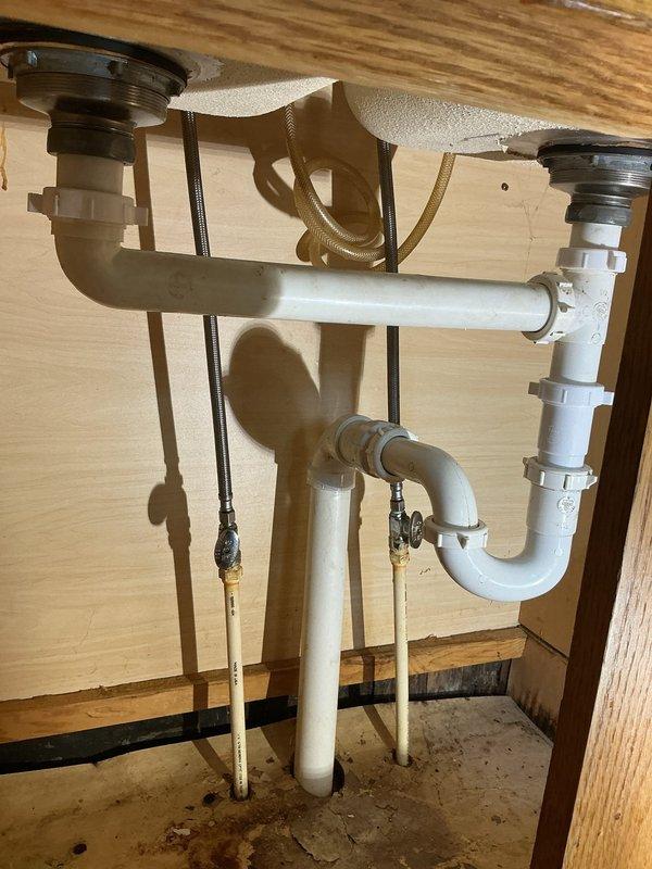 # Plumbing Service Completed

Responded to service call for leaking pipes under kitchen sink. Upon inspection, identified corroded drain pipes in double-basin sink configuration causing active water leakage. The existing white PVC drainage system showed signs of deterioration with visible corrosion at connection points beneath the cabinet.

Assessed the complete under-sink plumbing assembly, including dual P-trap system, horizontal drain connections, and supply lines. Replaced corroded pipe sections and reinstalled drainage components to ensure proper water flow and eliminate leaks. Verified all connections were secure and tested system for proper drainage and seal integrity. Cabinet interior showed water staining from previous leakage, confirming the need for repair.

System restored to full working condition with no active leaks detected upon completion.