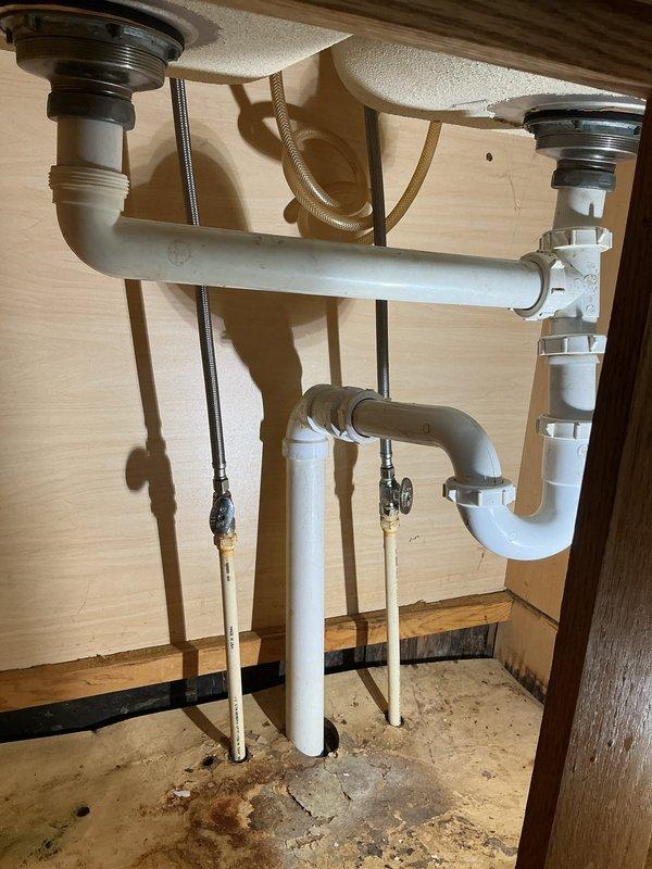 # Plumbing Service Completed

Responded to service call for leaking pipes under kitchen sink. Upon inspection, identified corroded drain pipes in double-basin sink configuration causing active water leakage. The existing white PVC drainage system showed signs of deterioration with visible corrosion at connection points beneath the cabinet.

Assessed the complete under-sink plumbing assembly, including dual P-trap system, horizontal drain connections, and supply lines. Replaced corroded pipe sections and reinstalled drainage components to ensure proper water flow and eliminate leaks. Verified all connections were secure and tested system for proper drainage and seal integrity. Cabinet interior showed water staining from previous leakage, confirming the need for repair.

System restored to full working condition with no active leaks detected upon completion.