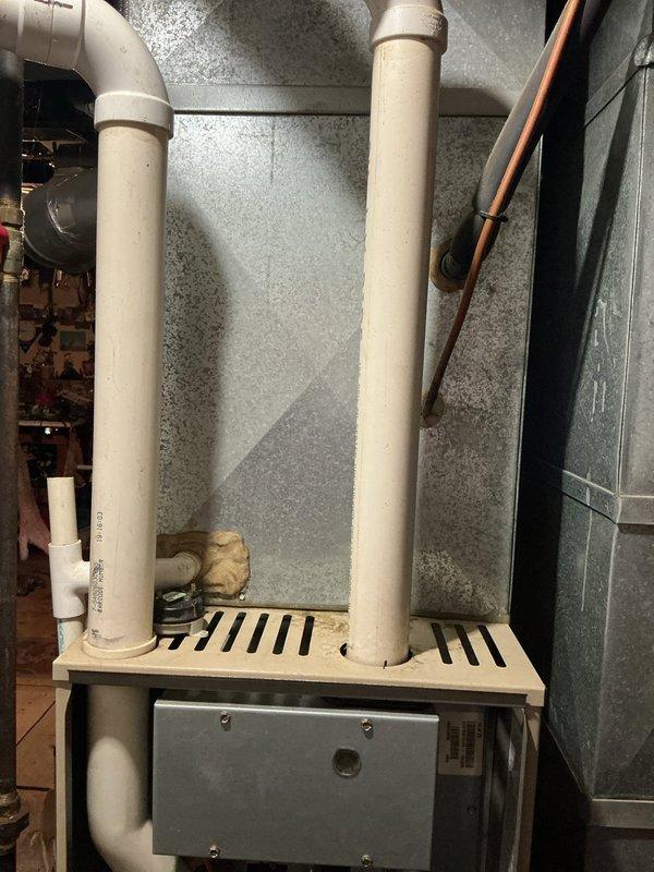 Completed furnace retrofit installation in residential basement utility room. Replaced existing heating unit with new furnace system, including proper venting configuration using PVC exhaust and intake piping routed through wooden platform support structure. Verified all connections to existing ductwork and ensured proper clearances from water heater and surrounding utilities. Exterior condenser unit remains in service on concrete pad adjacent to building exterior with stone veneer siding, connected via refrigerant lines for cooling operation. System tested and operational upon completion.