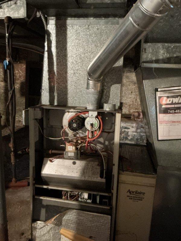 Completed furnace retrofit installation of Aprilaire HVAC system in basement utility area. Unit was properly positioned with access panels opened for final inspection of blower motor assembly, electrical connections, and control mechanisms. Verified all internal components including red motor wiring, mounted controls, and metallic vent piping extending from unit top are secured and functioning per manufacturer specifications.