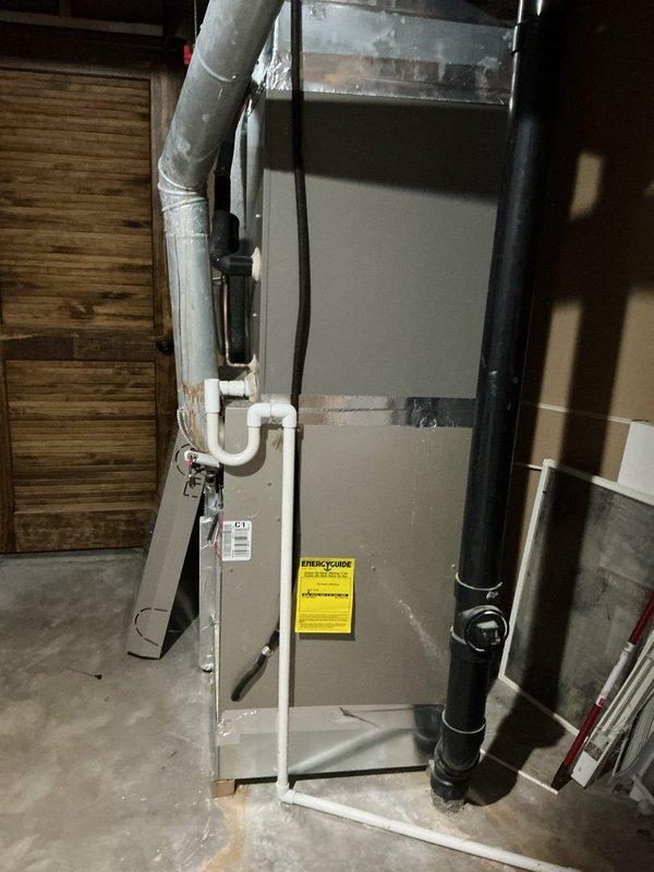 Completed furnace retrofit installation in basement utility area. New gray metal cabinet furnace unit installed against wall with proper PVC condensate drainage system, white insulated supply ductwork, and black flexible exhaust piping secured with metal clamps. All components properly connected and positioned for optimal operation in accordance with manufacturer specifications.