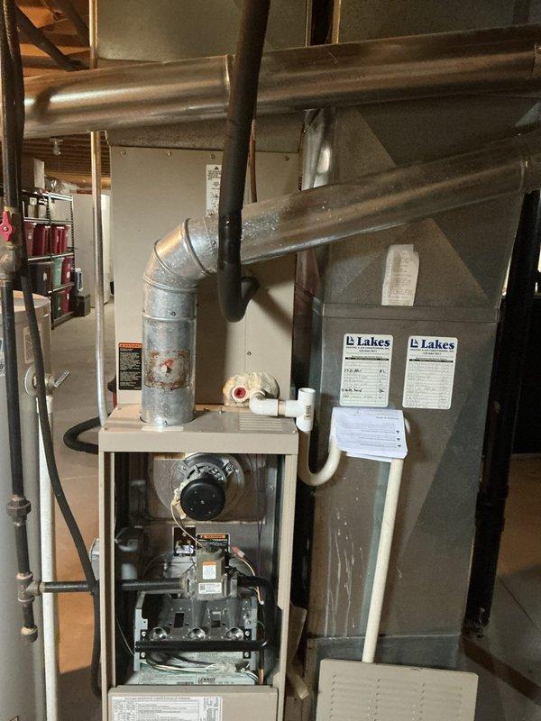 Completed furnace retrofit in residential basement mechanical room, documenting existing HVAC system configuration including furnace unit with exposed internal components, gas valve assembly, and control mechanisms. Photographed galvanized metal ductwork connections, PVC condensate piping, and adjacent Lakes-branded air handler unit for replacement planning documentation. Also documented existing Bradford White 40-gallon natural gas water heater (Model MI40S6FBN, 40,000 BTU/hr input) with all specification labels and safety warnings visible for comprehensive system assessment.