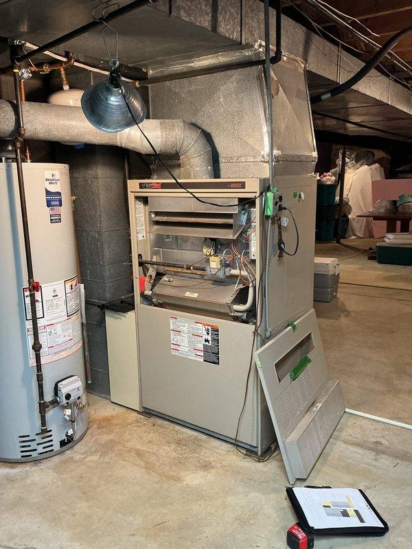 Performed an on-site evaluation for a multi-system retrofit replacement, inspecting the basement gas furnace with its burner assembly, gas valve, and wiring exposed, along with the adjacent water heater and existing supply plenum and flexible ductwork. Assessed the two exterior condenser units, noting the elevated unit mounted on blocks with debris accumulation and weathered cabinet panels, and documented the electrical disconnect and conduit configuration. Measured the equipment locations and took photographs to size and specify replacement furnace and condenser equipment.