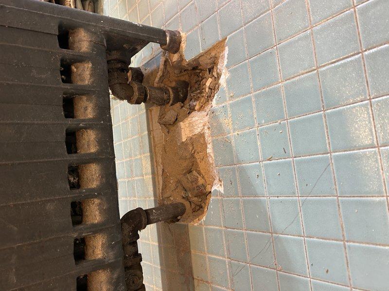 Performed diagnostic inspection of radiator system, documenting significant wall deterioration and moisture damage at pipe connection points. Temporarily removed radiator unit from damaged blue-tiled wall to assess extent of compromised mounting area and pipe fittings requiring future repair.