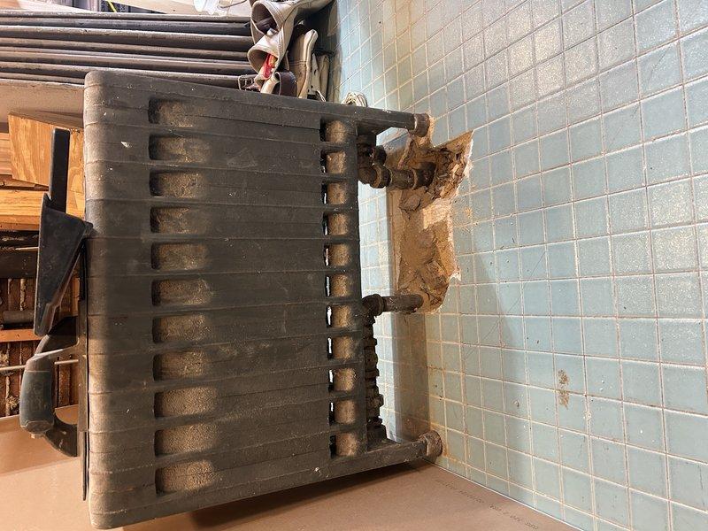 Performed diagnostic inspection of radiator system, documenting significant wall deterioration and moisture damage at pipe connection points. Temporarily removed radiator unit from damaged blue-tiled wall to assess extent of compromised mounting area and pipe fittings requiring future repair.