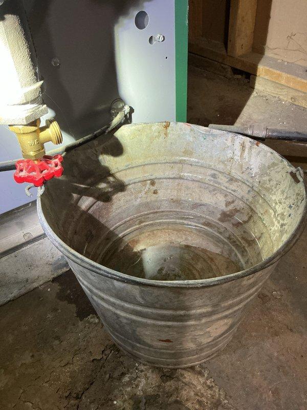 Responded to boiler leak complaint and performed diagnostic inspection. Upon arrival, found active water seepage from boiler system requiring immediate attention. Observed pressure gauge reading at zero position and noted brass drain valve assembly with collection bucket in place beneath unit showing signs of mineral deposits and water discharge. Identified leak source at lower boiler connection point. Completed call-of-the-week (COTW) service evaluation, documented system conditions with photographic evidence, and provided assessment findings. Unit requires follow-up repair to address leaking component and restore proper system operation.