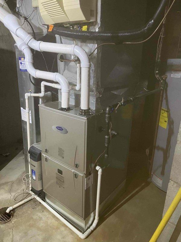 # HVAC Furnace Installation

Completed full furnace replacement in residential basement utility room. Removed existing heating unit and installed new Carrier brand vertical cabinet furnace on concrete floor pad. Configured all necessary venting with white PVC piping, including proper condensate drain lines with multiple elbows and connections routed upward and laterally for code-compliant exhaust. Connected black insulated refrigerant lines to existing air conditioning system. Integrated cream-colored air handler/humidifier unit with horizontal ventilation slats, mounted above furnace with appropriate plumbing connections. Wired all electrical connections to existing panel and verified proper voltage supply. Installed and connected wall-mounted thermostats for system control. Ensured proper clearances from adjacent water heater and other mechanical equipment. Tested all safety controls, ignition sequence, and airflow. System operational and heating efficiently.
