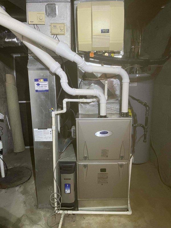 # HVAC Furnace Installation

Completed full furnace replacement in residential basement utility room. Removed existing heating unit and installed new Carrier brand vertical cabinet furnace on concrete floor pad. Configured all necessary venting with white PVC piping, including proper condensate drain lines with multiple elbows and connections routed upward and laterally for code-compliant exhaust. Connected black insulated refrigerant lines to existing air conditioning system. Integrated cream-colored air handler/humidifier unit with horizontal ventilation slats, mounted above furnace with appropriate plumbing connections. Wired all electrical connections to existing panel and verified proper voltage supply. Installed and connected wall-mounted thermostats for system control. Ensured proper clearances from adjacent water heater and other mechanical equipment. Tested all safety controls, ignition sequence, and airflow. System operational and heating efficiently.