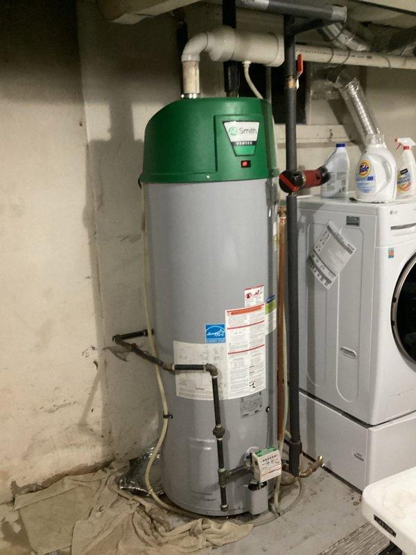 Responded to emergency service call for leaking 20-year-old A.O. Smith tanked water heater located in basement utility area. Upon arrival, conducted visual inspection of unit and surrounding plumbing connections, documenting existing conditions with photographs. Assessed water heater showing signs of age-related deterioration, multiple pipe connections including copper supply lines, PVC venting, pressure relief valve with discharge piping, and various shut-off valves. Unit displayed manufacturer specifications and energy compliance labels on exterior tank body with characteristic green crown assembly.