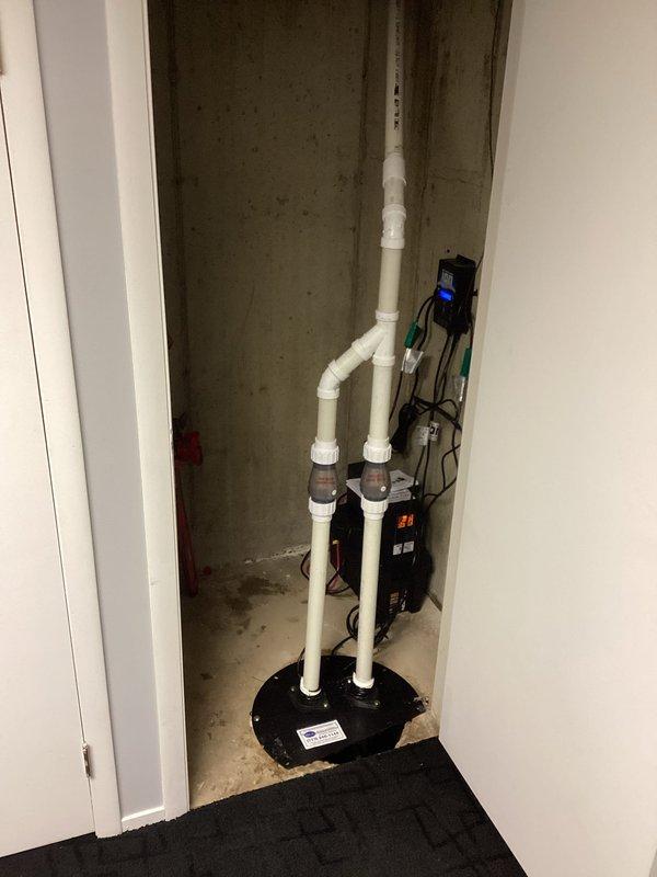 Completed installation of battery backup sump pump system with iOn Genesis control unit. System installed in utility closet with dual PVC discharge pipes running vertically from floor-mounted basin, connected via couplings to horizontal discharge line. Control panel mounted on wall with digital display for system monitoring and multiple cable connections to pump components secured with terminal blocks.