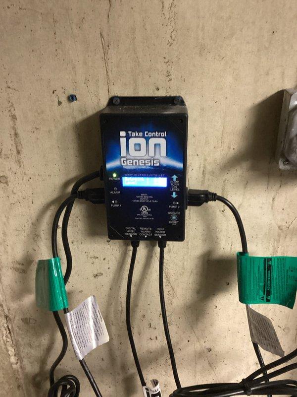 Completed installation of battery backup sump pump system with iOn Genesis control unit. System installed in utility closet with dual PVC discharge pipes running vertically from floor-mounted basin, connected via couplings to horizontal discharge line. Control panel mounted on wall with digital display for system monitoring and multiple cable connections to pump components secured with terminal blocks.