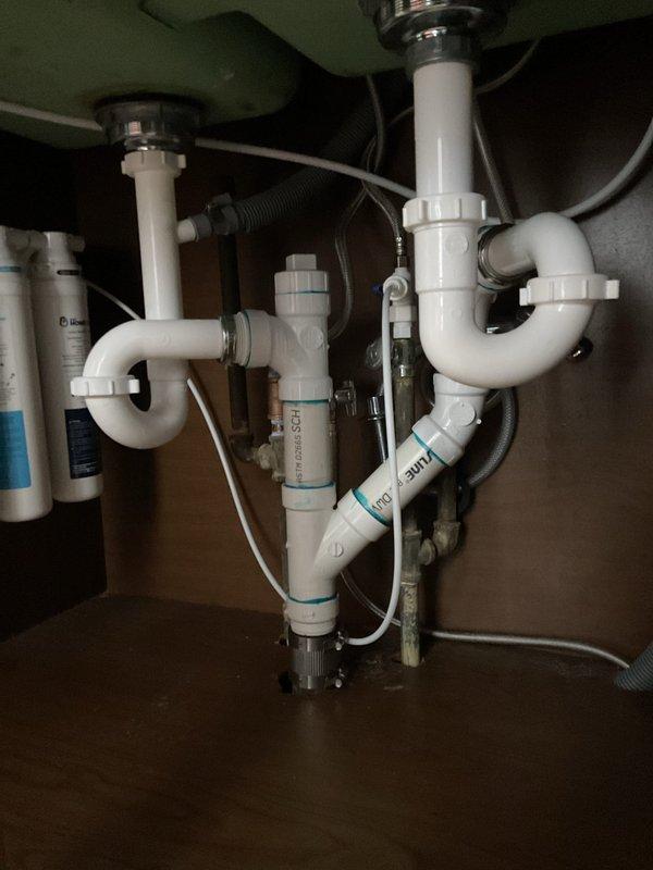 Responded to kitchen sink clog at residential property. Upon arrival, accessed cabinet beneath dual-basin sink to assess drainage system. Inspected white PVC P-trap assembly connected to chrome tailpieces and main vertical drain line. Cleared obstruction from affected drain line, restoring proper water flow and drainage function. Verified both sink basins draining correctly upon completion. System tested and returned to normal operation.
