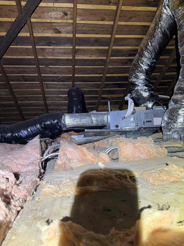 Conducted comprehensive HVAC system evaluation at residential property for replacement estimate of underperforming 3-year-old forced air system. Accessed attic space via ladder to inspect air handler unit, flexible ductwork configuration, and insulation coverage. Documented existing gray metal air handler installation with connected black insulated and metallic flexible ducting, noting duct routing patterns and connection points. Assessed fiberglass insulation condition, observing uneven coverage and gaps in both yellow and pink batt materials across attic floor joists. Photographed complete system layout including exposed wooden roof structure, diagonal support beams, and HVAC component placement for replacement planning purposes. Verified system accessibility and spatial constraints for future equipment upgrade considerations.