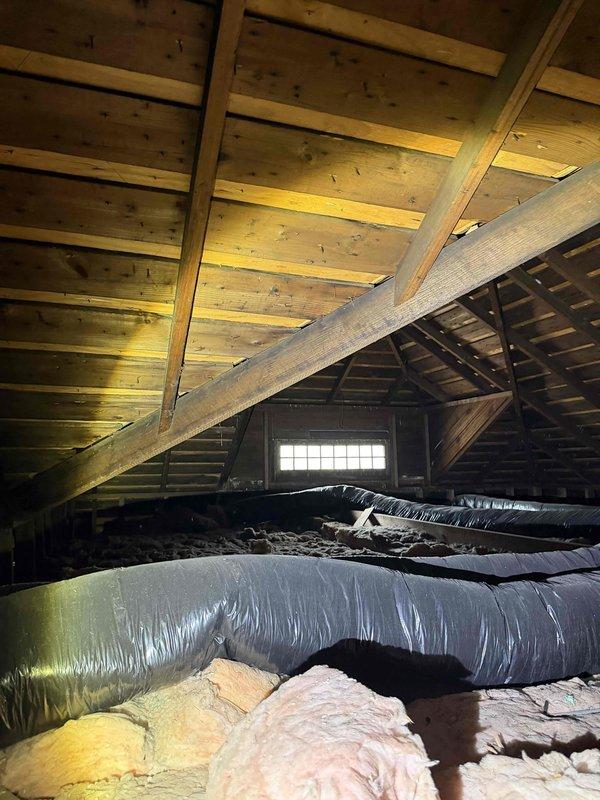 Conducted comprehensive HVAC system evaluation at residential property for replacement estimate of underperforming 3-year-old forced air system. Accessed attic space via ladder to inspect air handler unit, flexible ductwork configuration, and insulation coverage. Documented existing gray metal air handler installation with connected black insulated and metallic flexible ducting, noting duct routing patterns and connection points. Assessed fiberglass insulation condition, observing uneven coverage and gaps in both yellow and pink batt materials across attic floor joists. Photographed complete system layout including exposed wooden roof structure, diagonal support beams, and HVAC component placement for replacement planning purposes. Verified system accessibility and spatial constraints for future equipment upgrade considerations.