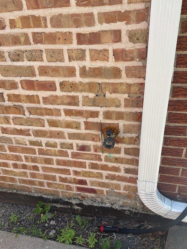 Completed inspection of 8-year-old frost-free hose bib requiring replacement at residential property. Examined exterior wall-mounted fixtures on red brick facade and assessed interior plumbing access point. Documented existing hose bib condition, wall penetration location, and surrounding infrastructure including downspout proximity to replacement area.