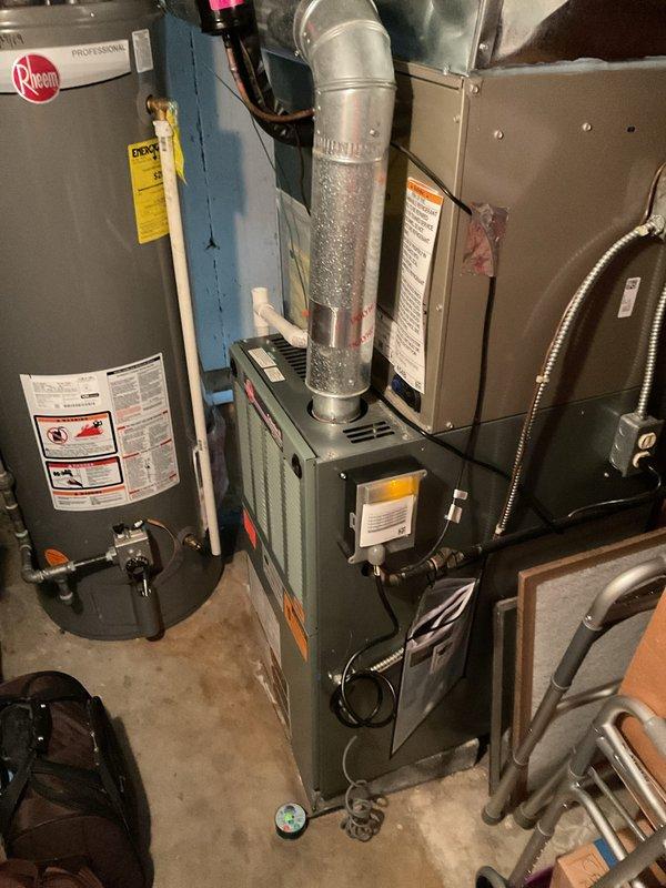 Responded to warranty call for non-operational thermostat following recent AC installation. Upon arrival, found thermostat completely removed from wall with exposed wiring at ceiling location. Documented existing conditions showing brown, blue, and yellow/green striped wires protruding from opening with visible drywall patching around fixture area. Inspected HVAC system in mechanical room, verifying furnace and ductwork connections were intact. Photographed equipment layout including condensation on vertical duct run. Thermostat requires replacement to restore system operation.