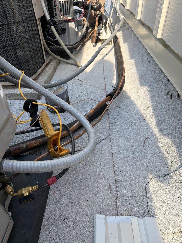 Relocated rooftop condenser unit with new refrigerant line routing across flat membrane roof surface. Secured copper refrigerant lines with metal straps and yellow plastic brackets, running insulated and bare copper piping between equipment locations. Electrical relocation work was not included in this scope of work.
