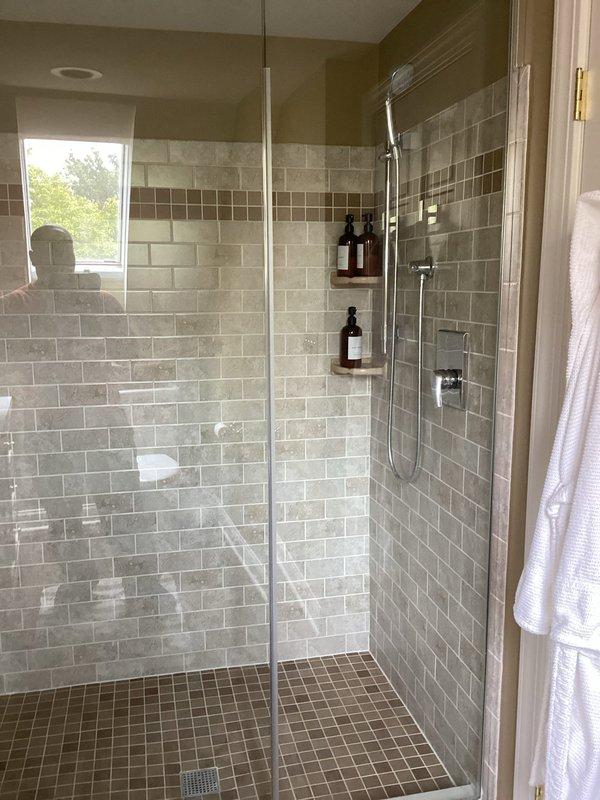 Completed comprehensive plumbing inspection of residential property, examining two full bathrooms for proper fixture operation, drainage, and potential issues. Inspected modern walk-in shower with glass enclosure, verifying chrome handheld showerhead functionality, sliding rail mounting, shower controls operation, and proper drainage through floor-mounted square drain; confirmed subway tile and mosaic accent installations showed no signs of water intrusion or grout deterioration. Evaluated second bathroom featuring vintage-style fixtures with brass/gold-toned components, including shower enclosure with glass doors and brass hardware, toilet operation, and wooden vanity with marble countertop and gold-toned faucet; verified all fixtures operated correctly with adequate water pressure and proper drainage throughout both bathrooms with no leaks detected.