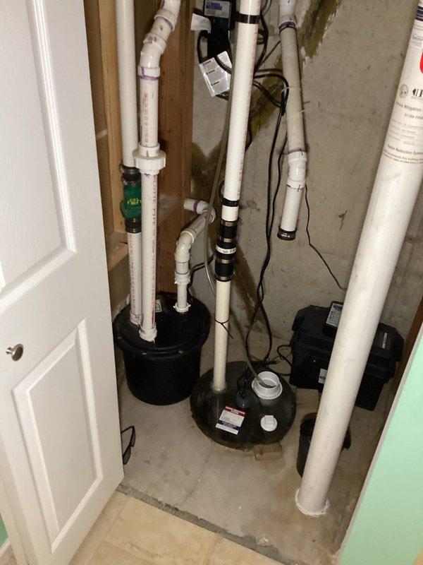 Conducted on-site assessment of existing sump pump and laundry ejector pit pump systems requiring replacement. Inspected utility room containing two black cylindrical pump units with associated white PVC discharge piping and electrical connections. Evaluated current Watchdog sump pump installation, noting vertical discharge pipe configuration, electrical wiring routing, and pump positioning within pit. Assessed laundry ejector pit pump system, examining discharge plumbing, vent piping, and power supply connections. Documented existing piping layout including various diameter PVC pipes, fittings, elbows, and connection points throughout the mechanical space. Photographed current equipment configuration, pipe routing, and installation details for estimate preparation. Noted water staining on concrete walls indicating previous moisture exposure and verified proper drainage requirements for replacement equipment. Reviewed electrical supply connections and wire routing through wooden framin