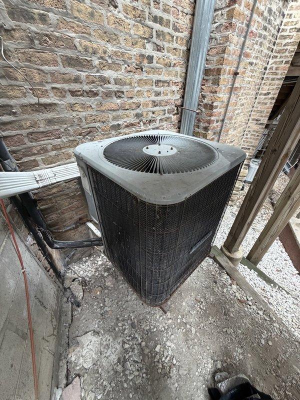 Responded to callback for AC unit noise complaint reported at previous service visit. Upon arrival, accessed unit located behind exterior steps via garage entry. Performed visual inspection and operational assessment of outdoor condenser unit positioned between brick walls on concrete pad. Unit appeared weathered with debris accumulation around base. Documented current condition of equipment, protective grille, and refrigerant lines with photographic evidence for service records.