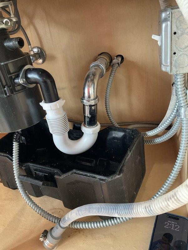 Completed plumbing inspection and diagnostic evaluation of kitchen sink system. Assessed all under-sink components including P-trap assembly, garbage disposal unit, supply lines, and drain connections. Documented existing installation featuring chrome P-trap with white threaded connectors, functional garbage disposal with discharge piping, and flexible water supply lines with proper fittings. Verified proper drainage function and photographed complete plumbing configuration for service records.