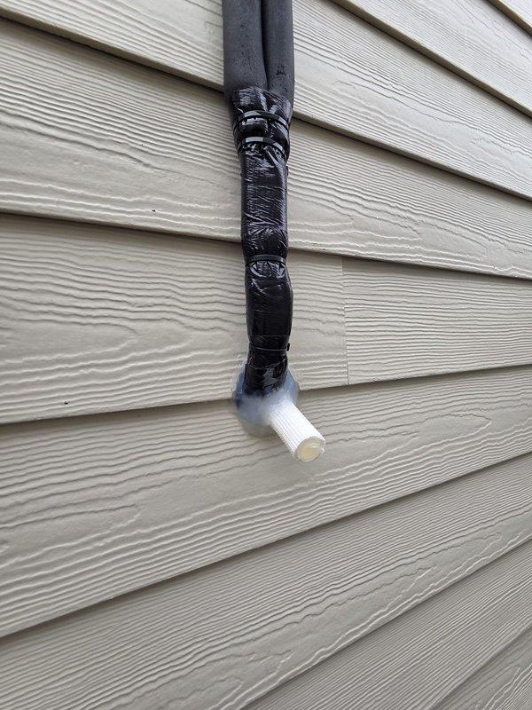 Completed site inspection and documented exterior drainage system condition. Observed black PVC condensate drain line with tape-wrapped sections running vertically along building exterior, terminating in white PVC elbow joint. Photographed drainage components and gutter system for service records.