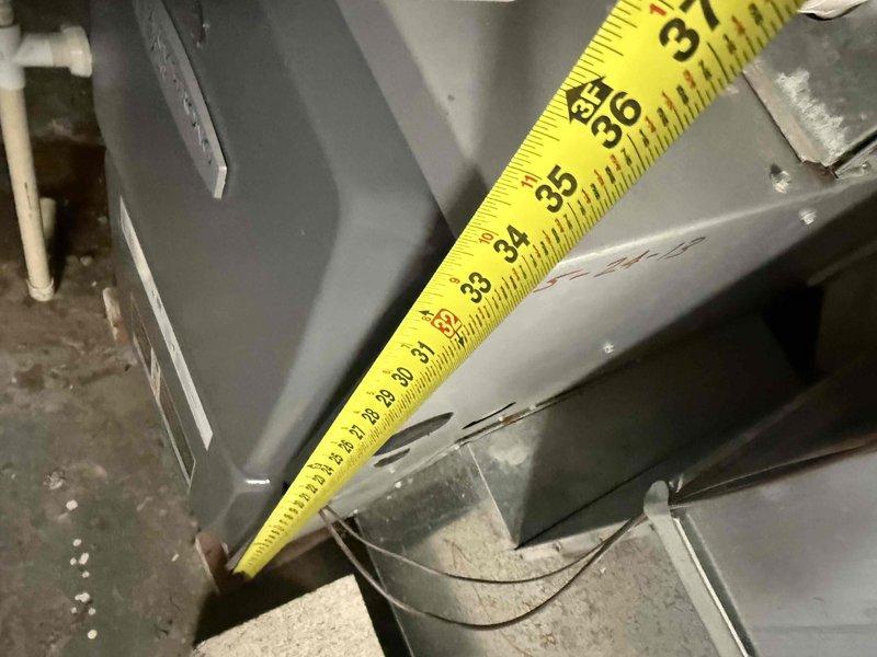 Conducted comprehensive assessment of 18+ year old furnace system experiencing inadequate cooling performance. Documented existing basement-installed HVAC unit with dark gray cabinet, PVC venting, and associated ductwork configuration. Performed detailed measurements of equipment dimensions and clearances using precision measuring tools. Evaluated complete system including furnace cabinet, supply and return ductwork, venting components, and installation environment. Photographed equipment nameplate data, unit positioning, ductwork connections, and mechanical room layout to develop accurate replacement specifications for full system estimate.