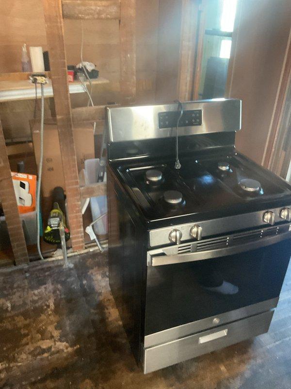 Completed gas range hookup and installation in residential property undergoing renovation with extensive fire or water damage to subfloor. Connected gas line to freestanding four-burner range with stainless steel controls in unfinished space with exposed framing, ensuring proper ventilation and code-compliant installation despite challenging site conditions.