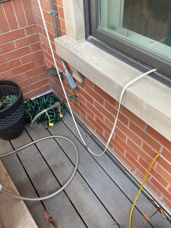 Conducted on-site evaluation for natural gas line modification to accommodate new pizza oven installation on outdoor patio area. Assessed existing gas hookup currently serving grill to determine feasibility of adding T-connector for dual appliance configuration. Documented copper gas line penetrations through brick wall, existing plumbing connections, and available workspace on composite deck surface for proposed installation work.
