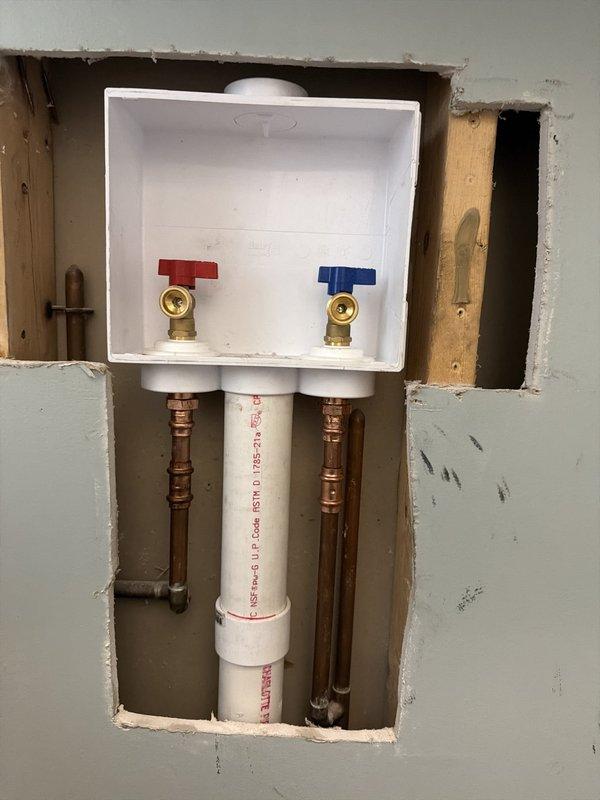 Replaced existing grey laundry box with new white Oatey outlet box assembly, installing two shut-off valves with quarter-turn handles (red and blue) connected to existing copper supply lines and PEX tubing. Installation completed with black flexible supply lines extending from mounted valves, providing proper water supply connections for washing machine hookup.