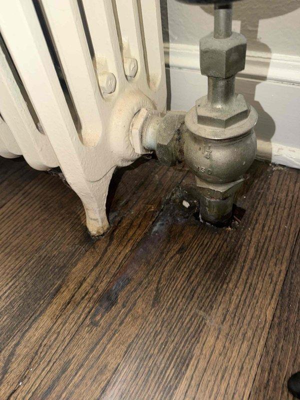 Responded to report of water leaking from first-floor radiator in home with 16-year-old boiler system. Upon inspection, identified severe corrosion on radiator valve assembly and associated piping connections. The thermostatic radiator valve (TRV) and metal elbow joint showed significant deterioration with green and yellowish-brown oxidation, crusty deposits, and compromised integrity at connection points. Evidence of long-term moisture exposure was apparent with rust stains and water damage on surrounding surfaces. Repaired the leaking valve assembly, replaced corroded components, and verified system integrity to restore proper heating function and eliminate active water leak.