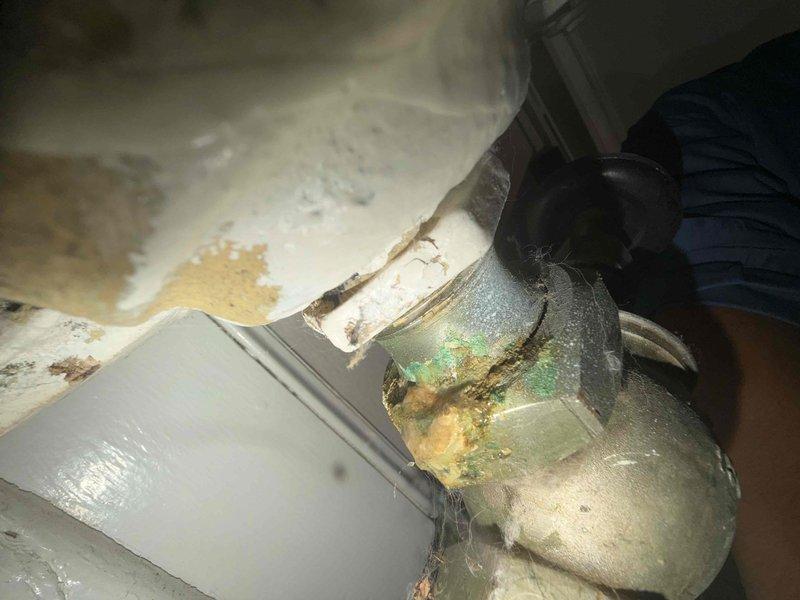 Responded to report of water leaking from first-floor radiator in home with 16-year-old boiler system. Upon inspection, identified severe corrosion on radiator valve assembly and associated piping connections. The thermostatic radiator valve (TRV) and metal elbow joint showed significant deterioration with green and yellowish-brown oxidation, crusty deposits, and compromised integrity at connection points. Evidence of long-term moisture exposure was apparent with rust stains and water damage on surrounding surfaces. Repaired the leaking valve assembly, replaced corroded components, and verified system integrity to restore proper heating function and eliminate active water leak.