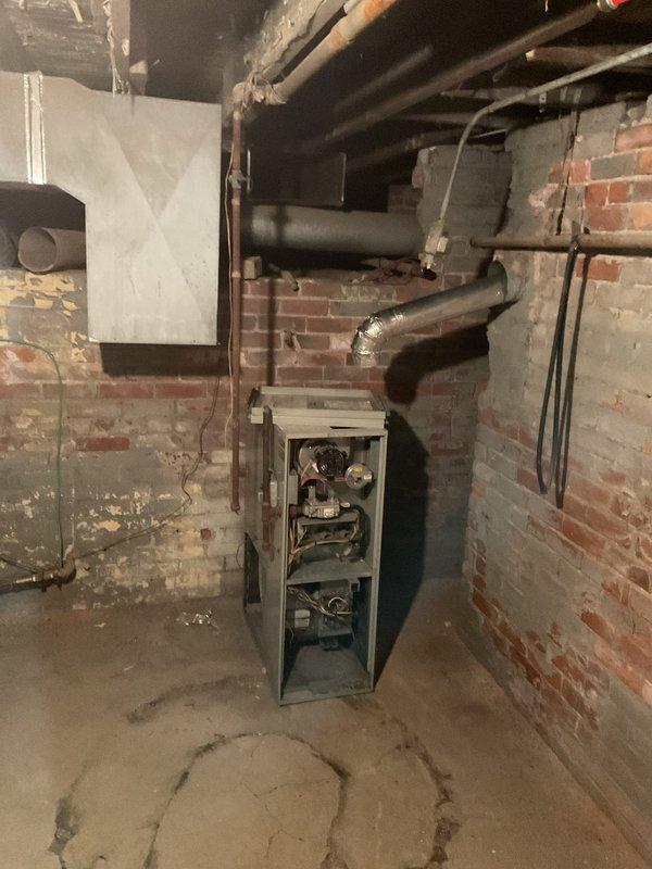 Performed comprehensive assessment of two aging furnaces located in basement utility area with exposed brick walls and deteriorating infrastructure. Evaluation revealed outdated units with accessible internal components requiring complete replacement. Documented existing ductwork configuration, including rectangular sheet metal junction and connecting pipes, to ensure proper sizing and compatibility with replacement systems.