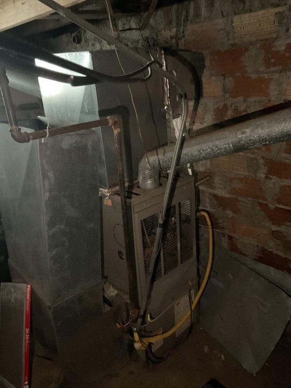 Performed inspection of existing furnace system in basement utility area. Unit shows signs of age and wear with deteriorating components. Evaluated ductwork condition, noting metal connections along concrete ceiling. Documented current installation against brick wall with copper piping and electrical connections. Provided replacement options for complete system upgrade based on assessment.