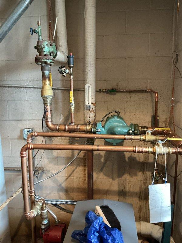 Performed diagnostic assessment on 10+ year old boiler system that was non-operational. Inspected complex piping arrangement including pressure regulator, multiple valves, and copper distribution lines. Identified oxidation on central valve assembly and signs of water damage on surrounding concrete block wall; recommended system replacement due to age and deterioration of critical components.