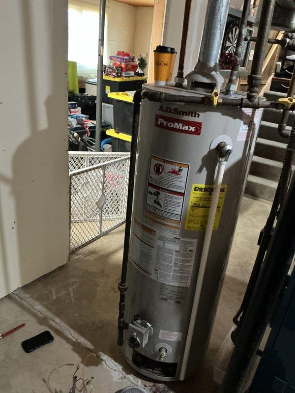 Responded to service call for reported tankless water heater malfunction at residential property. Upon arrival, found A.O. Smith ProMax conventional tank-style water heater installed in basement utility room, not a tankless unit as initially reported. Performed diagnostic inspection of water heater and associated plumbing connections, including copper supply lines, shut-off valves, and venting system. Verified proper installation configuration with metallic ductwork and exhaust venting. Unit appeared operational with all safety labels and manufacturer specifications visible and intact. Documented existing installation with photographs showing water heater positioning, piping connections, and mechanical room layout for service records.