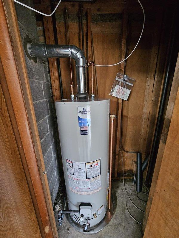 Completed installation of a 50-gallon Bradford White tanked water heater in residential basement utility area. Drained and removed existing water heater unit, then positioned new water heater with proper drain pan for code compliance and leak protection. Upsized ventilation flue from existing diameter to 4-inch configuration, extending through concrete block wall to chimney connection with proper sealing and mortar application at wall penetration point. Replaced old piercing valve with new valve for icemaker water supply line to ensure reliable connection and prevent future leaks. Removed flexible gas connector and installed hard-piped gas line using black iron pipe with appropriate fittings for enhanced safety and code compliance. Connected all copper water supply lines at top of unit with proper metallic fittings and verified secure connections. Ensured all plumbing connections, gas line, and ventilation components meet current building codes and manufacturer specifications for safe 