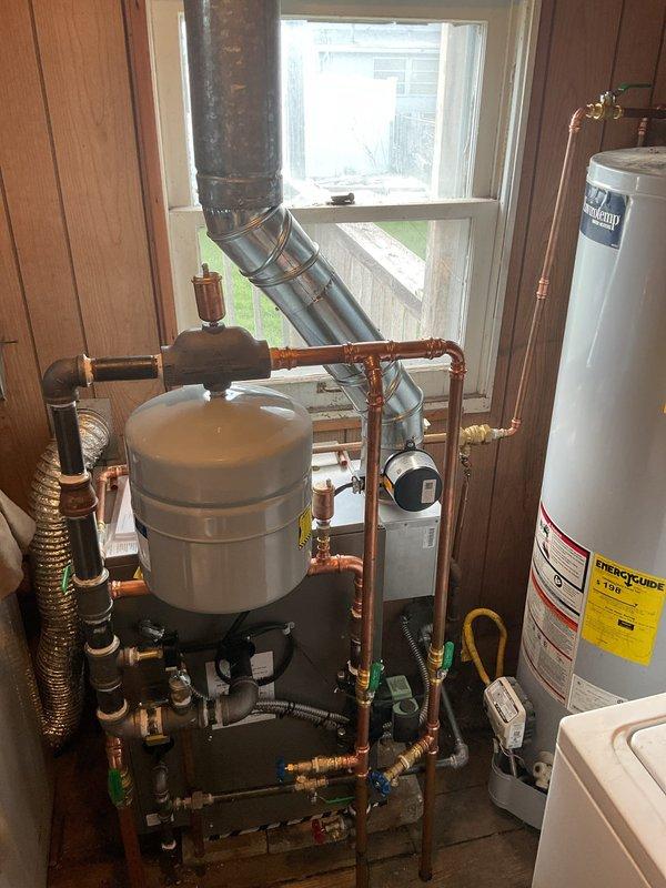 # HVAC Install - Boiler

Completed full boiler replacement in residential mechanical room. Removed existing boiler unit and mounted new boiler on stable, level surface with proper clearances. Connected supply and return piping to boiler using copper pipe with appropriate fittings, joints, and elbows. Installed required safety devices including pressure relief valve and water level gauge per manufacturer specifications and code requirements.

Connected gas line to main gas supply with secure, leak-free connections and installed gas shutoff valve for system safety. Performed complete electrical wiring per manufacturer's diagram and local electrical codes, connecting control panel, thermostats, and zone controls as required for proper system operation.

Installed white cylindrical expansion tank on metal frame with proper piping connections to accommodate system thermal expansion. Routed black vertical vent piping through ceiling for proper combustion exhaust. Integrated new boiler with e
