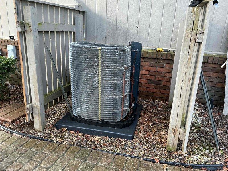 Completed second appointment to troubleshoot HVAC system with problematic dampers previously identified during initial diagnostic visit. Performed comprehensive A/C tune-up on outdoor condensing unit, including inspection of cylindrical coil assembly which showed weathering, mineral deposits, and debris accumulation between fins. Documented unit condition with exterior coil displaying surface discoloration and foam insulation material along refrigerant line connections, with system mounted on rubber pad in gravel bed adjacent to property fence line.