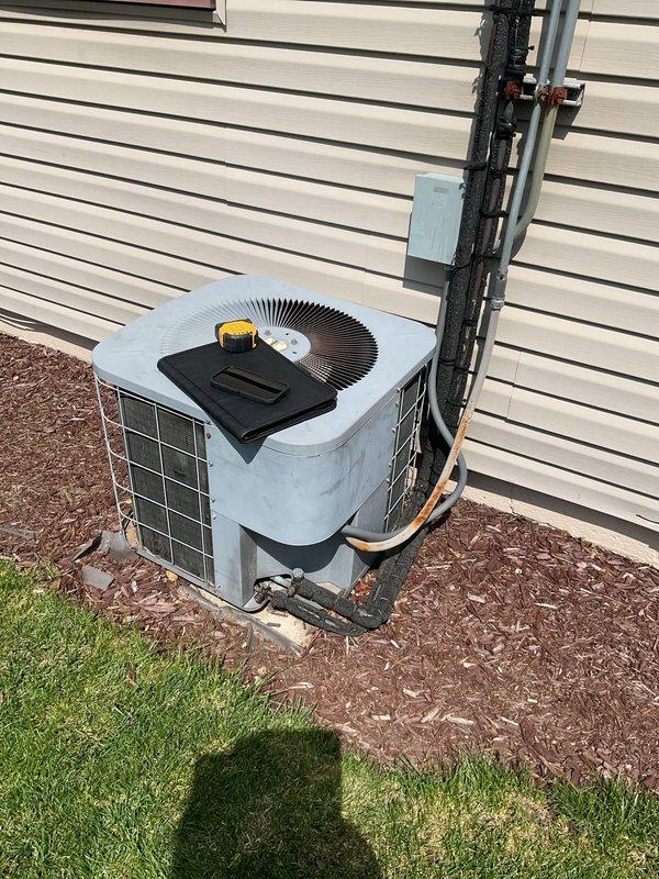 Conducted comprehensive AC system estimate for complete HVAC replacement at residential property. Evaluated existing outdoor condensing unit with gray/blue cabinet positioned on mulch bed against building exterior, noting refrigerant line routing along siding with proper insulation and mounting brackets. Assessed indoor gray metal furnace unit located in utility room with beige tile flooring, observing multiple ductwork configurations extending from unit top and flexible ductwork components. Documented current system configuration including refrigerant line connections, electrical wiring pathways, gas line positioning, and emergency shut-off switch placement. Photographed equipment conditions, installation specifications, and surrounding infrastructure to provide accurate replacement system sizing and installation planning recommendations.