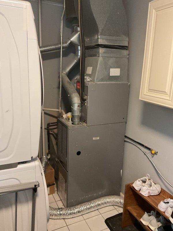 Conducted comprehensive AC system estimate for complete HVAC replacement at residential property. Evaluated existing outdoor condensing unit with gray/blue cabinet positioned on mulch bed against building exterior, noting refrigerant line routing along siding with proper insulation and mounting brackets. Assessed indoor gray metal furnace unit located in utility room with beige tile flooring, observing multiple ductwork configurations extending from unit top and flexible ductwork components. Documented current system configuration including refrigerant line connections, electrical wiring pathways, gas line positioning, and emergency shut-off switch placement. Photographed equipment conditions, installation specifications, and surrounding infrastructure to provide accurate replacement system sizing and installation planning recommendations.