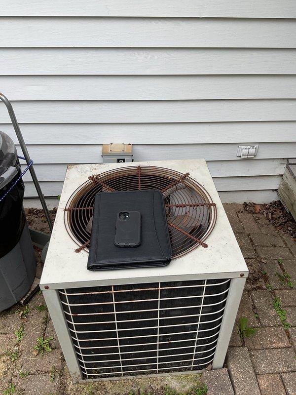 Conducted comprehensive site evaluation of existing outdoor air conditioning condenser unit for replacement estimate. Unit exhibited visible aging, wear, and discoloration consistent with extended service life. Documented current installation configuration including electrical disconnect box, conduit routing, refrigerant line sets with insulation sleeves, and equipment positioning on brick paver base. Assessed existing infrastructure conditions and photographed equipment specifications to determine appropriate replacement system requirements and installation parameters.